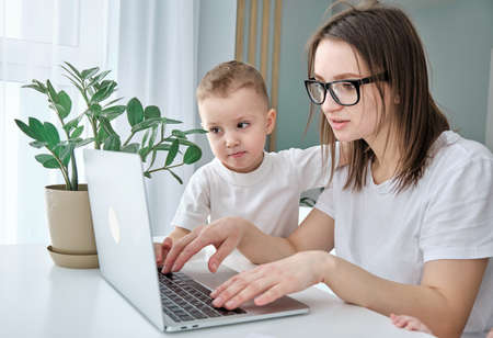 A young freelance mother works from home remotely using a laptop, her son sits nearby.の写真素材