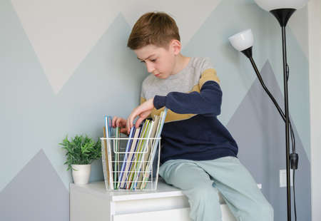 Young boy sitting and reading a book on a chest of drawers.