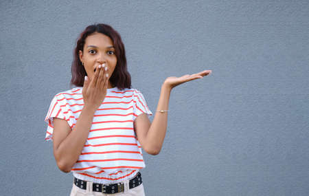 Amazed shocked Afro woman shows good sale offer or deal, holds breath, demonstrates empty copy space for promo text, surprised by big discounts, isolated over blue background. Advertisement.の写真素材