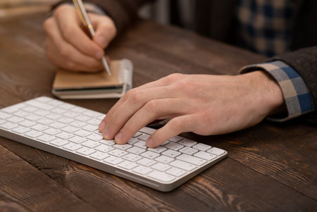 close-up of hands of person taking noted on paper white typing on computer keyboard
