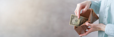 Young woman with sad expression holds almost empty brown leather wallet with few dollars, wearing...