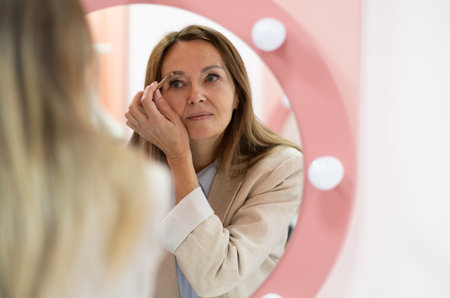Elegant senior woman applying blush with a makeup brush, focused on her reflection. She wears a...