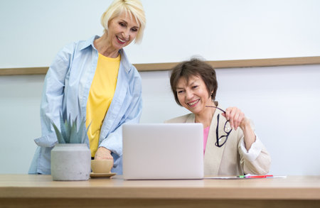 Two cheerful elderly female colleagues collaborating on a laptop in a modern office, surrounded by colorful charts. Smiling and engaged in a discussion, showcasing teamwork and productivity.の写真素材