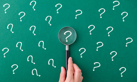 A hand holding a magnifying glass over a green background with many questions. Concept of curiosity and exploration, as if the person is trying to uncover the answers to these questionsの写真素材