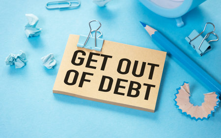 On a light blue background, a sticky note with text about debt management, alongside office supplies including paper clips and pencil shavingsの写真素材