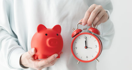 A person is holding a red piggy bank and a red alarm clock. The piggy bank is red and has a cute...