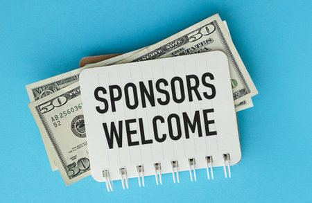 A stack of money is on top of a white sign that says Sponsors Welcome. The sign is on a blue backgroundの写真素材