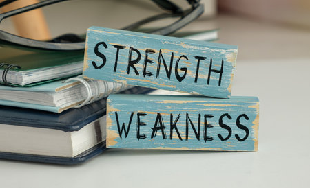 Two wooden blocks with the words strength and weakness written on them. The blocks are placed on top of a stack of books, which are also on a table. Concept of balance and contrastの写真素材