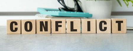 A wooden block with the word Conflict written on it. The block is placed on a table, and there are other items on the table, such as a calculator, a book, and a potted plantの写真素材