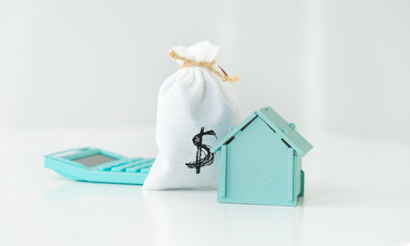 A blue house is next to a calculator and a bag of money. The calculator is on a table and the bag of money is on the floorの写真素材