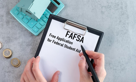 A person is writing on a piece of paper that says FASFA on it. They are using a pen to write on the paper. The paper is placed on a table, and there is a calculator nearbyの写真素材