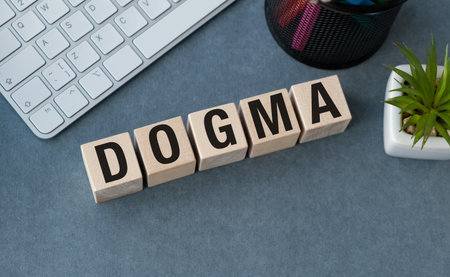 A wooden block with the letters DOGMA written on it. The block is on a desk with a keyboard and a potted plantの写真素材