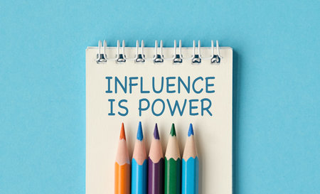 A notebook with a spiral binding and a pencils on top of it. The pencils are in different colors and are arranged in a row. The notebook has the words Influence is Power written on itの写真素材