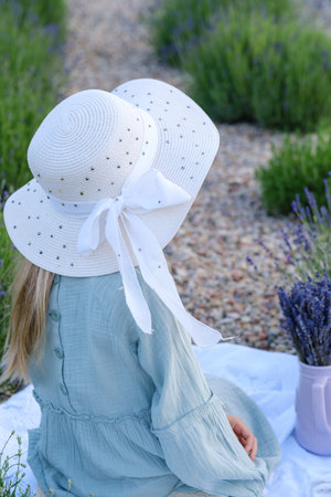A girl wearing a white hat and a blue dress is sitting on a blanket. There are purple flowers in a vase nearbyの写真素材