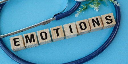 A stethoscope is on a blue background with the word Emotions written in wooden blocks. The stethoscope is on the left side of the imageの写真素材