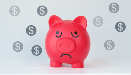 A red piggy bank with a sad face and a pile of dollar coins. The piggy bank is empty, and it seems to be feeling downの写真素材