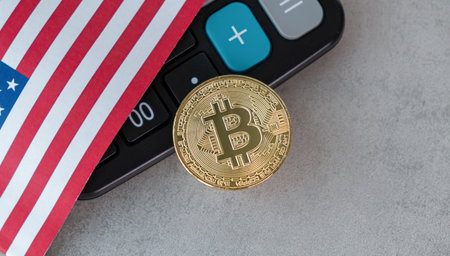 A calculator is on a table with a coin that says Bitcoin on it. The calculator is on top of a piece of paper with the American flag on itの写真素材