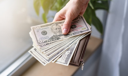 A person is holding a wallet full of money. The wallet is brown and has a zipper