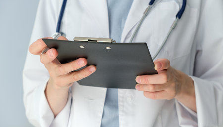A doctor is holding a clipboard and writing on it. The clipboard is black and white. The doctor is wearing a white coatの写真素材