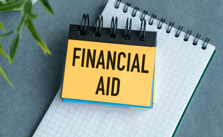 A yellow sticky note with the word Financial Aid written on it. The note is sitting on top of a spiral bound notebookの写真素材