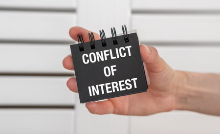 A hand holding a notebook with the word Conflict of Interest written on it. The notebook is black...