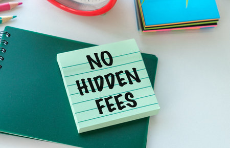 A green notebook with a white sticker that says No Hidden Fees. The notebook is on a table with other items such as a pencil, a pen, and a clockの写真素材