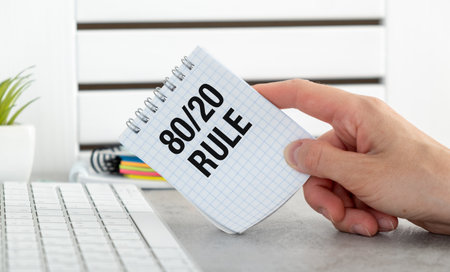 A person is holding a piece of paper that says 80 and 20 rule. The paper is on a desk next to a...