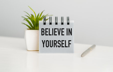 A small plant sits on a table next to a notebook that says Believe in Yourself. A pen is also on the...