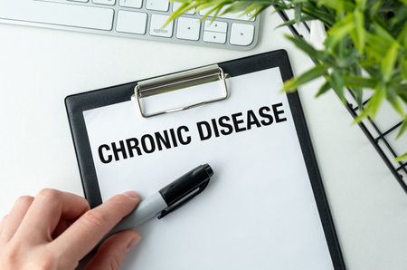A person is writing the word Chronic Disease on a clipboard. The clipboard is on a table with a keyboard and a plant in the backgroundの写真素材