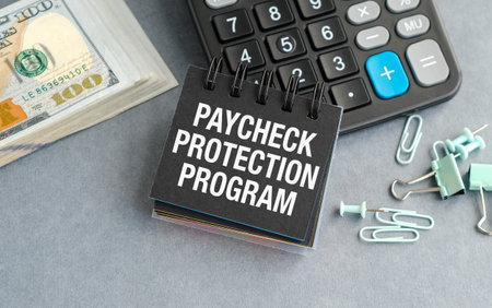 A calculator sits on a desk with a stack of money and a piece of paper that says Paycheck Protection Programの写真素材