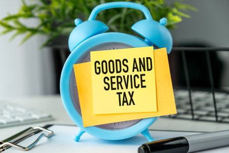 A blue alarm clock with a yellow note on it that says Goods and Service Tax. The clock is placed on a table with a pen and a keyboardの写真素材
