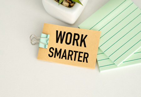 A brown tag with the Word Work Smarter on it. It is hanging from a clip. There is a green plant in the backgroundの写真素材