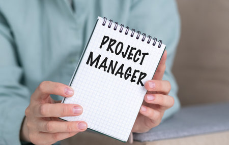 A person holding a notebook with the word Project Manager written on it. The notebook is open to a page with the word project manager written in black markerの写真素材