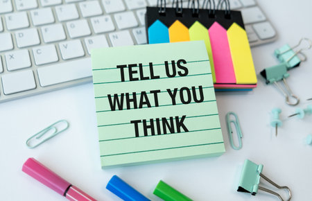 A green and white sign that says Tell us What you Think is on a desk with a keyboard and a stack of...