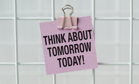 A pink piece of paper with the words Think About Tomorrow Today written on it. The paper is clipped to a white metal fenceの写真素材