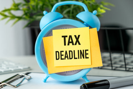 A blue alarm clock with a yellow note on it that says Tax Deadline. The clock is on a table with a keyboard and a penの写真素材