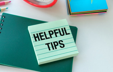 A green notebook with a white sticker that says Helpful Tips on it. The notebook is sitting on a table with other items such as a pencil, a pen, and a clockの写真素材