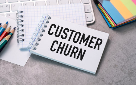 A white spiral notebook with the word customer churn written on it. The notebook is on a desk with a keyboard and a pencilの写真素材