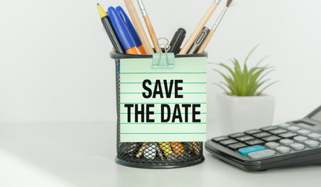 A pen holder with a green and white sign that says Save the Date. The pen holder is on a table next to a calculatorの写真素材