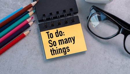 A yellow sticky note with the words To Do: So Many Things written on it. The note is placed on a desk next to a pair of glasses and a box of colored pencilsの写真素材