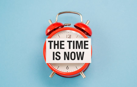 A red alarm clock with a white sign that says "The time is now.". The clock is set to the time of 10:30の写真素材