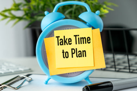 A blue alarm clock with a yellow note that says Take Time to Plan. The clock is placed on a desk...