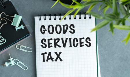 A notebook with the words Goods Services Tax written on it. The notebook is on a table with a pair of scissors and a pair of green paper clipsの写真素材