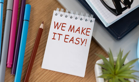A notebook with the words We Make it Easy written on it. The notebook is on a desk with a pencil, pens, and a laptopの写真素材