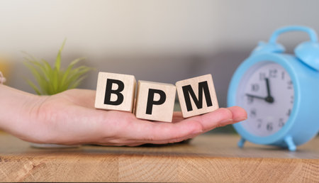 A hand holding three wooden blocks that spell out BPM. The blocks are on a table next to a blue clockの写真素材