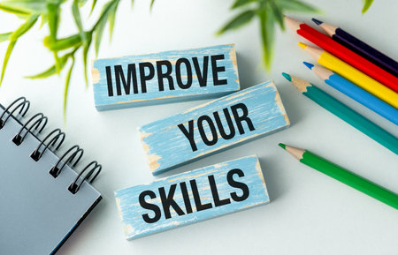A stack of wooden blocks with the words Improve Your Skills written on them. The blocks are placed on a table next to a notebook and a set of colored pencils. Concept of personal growthの写真素材