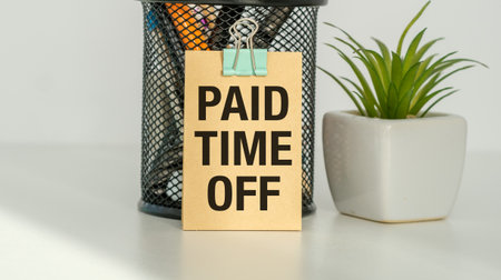 A small sign that says Paid Time Off is sitting on a desk. The sign is attached to a metal clip and is surrounded by pens and pencils. The desk is also topped with a potted plantの写真素材