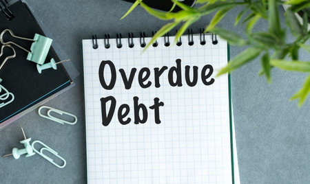 A notebook with the word Overdue Debt written on it. The notebook is on a table with a pair of...