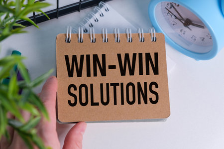 A person is holding a notebook with the words Win-Win Solutions written on it. The notebook is on a table next to a clockの写真素材