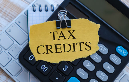 A calculator sits on a desk with a piece of paper that says Tax Credits. The calculator is black and silver, and the paper is yellow and has a clip holding it in placeの写真素材